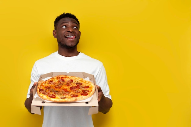 Um homem afro-americano alegre de camisa branca com uma caixa de pizza a lamber os lábios e a sonhar.