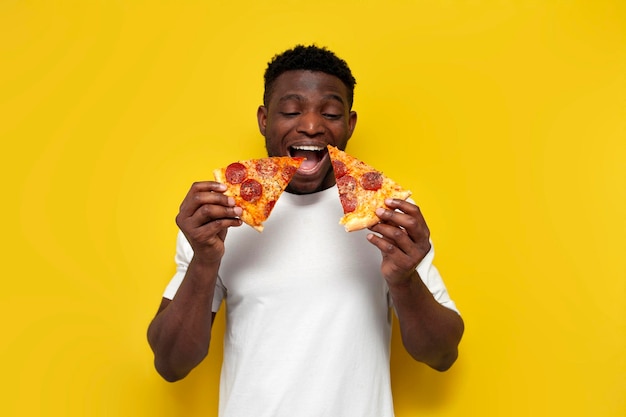 Um homem afro-americano alegre de camisa branca come duas fatias de pizza com a boca aberta.