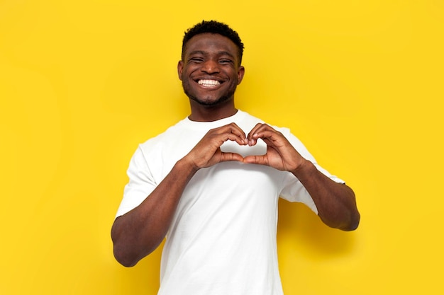 Um homem afro-americano alegre de camisa branca mostra o coração com as mãos em fundo amarelo