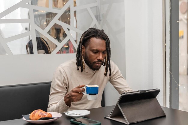 Um homem afro-americano bonito sentado na cafeteria com uma xícara de café usando um tablet digital