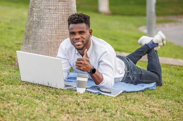 um homem afro-americano deitado na grama verde sorrindo e polegares para cima trabalhando em um computador portátil