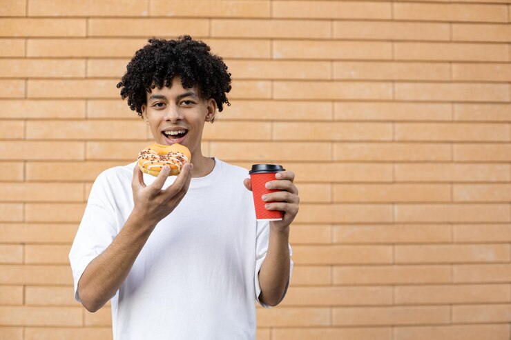 Foto um homem afro-americano feliz mordendo um donut laranja e segurando uma xícara de café