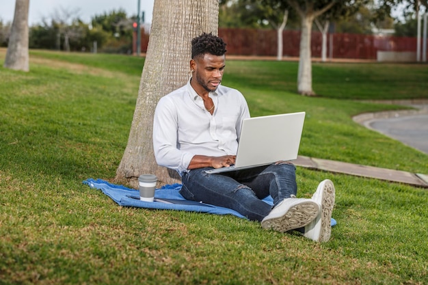 Um homem afro-americano senta-se debaixo de uma árvore trabalhando em seu computador portátil