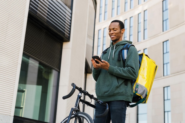 Um homem afro-americano sorridente durante a entrega de pizza no escritório O homem olha para a tela do telefone Entrega rápida de bicicleta