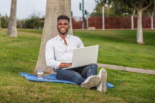 Um homem afro-americano sorridente sentado sob uma árvore trabalhando ou estudando com um computador portátil