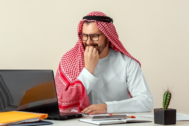 Um homem árabe, um empresário, um xeque trabalha em um laptop. Investimentos, negócios, trabalho pela Internet, contratos online.