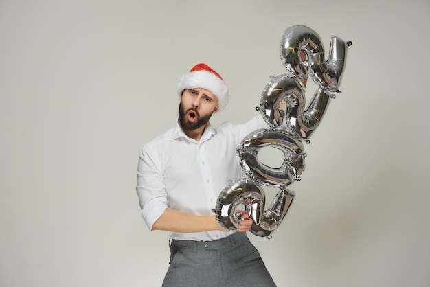 Um homem arrojado com um chapéu de Papai Noel de veludo vermelho com os lábios em um tubo empurra balões prateados no formato de 2022 na vertical. Um cara com barba em uma festa de ano novo.