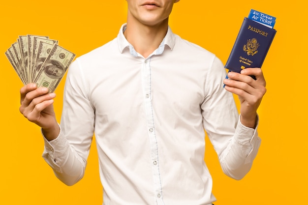 Um homem asiático bonito em uma camisa branca se alegra em ganhar na loteria. Ele está segurando um passaporte com passagens aéreas e dólares em um fundo amarelo. - imagem