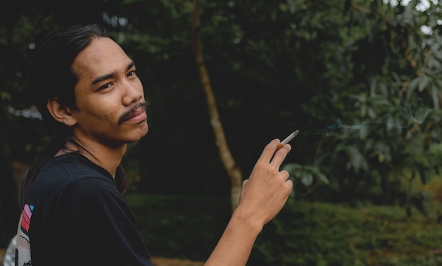 Um homem asiático de cabelos compridos com barba segurando um cigarro do lado de fora.