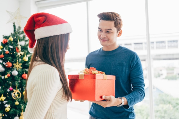 Um homem asiático feliz surpreende sua namorada com presentes de Natal em casa com uma árvore de Natal na imagem de fundo com espaço para cópia