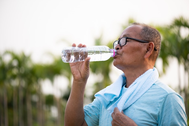 Um homem asiático velho feliz é água potável no parque com luz solar