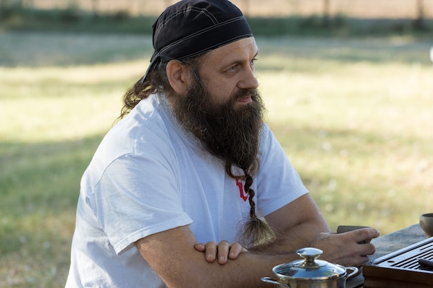 Um homem barbudo com uma bandana está bebendo chá no fundo da natureza.