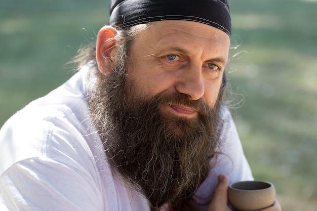 Um homem barbudo com uma bandana está bebendo chá no fundo da natureza.
