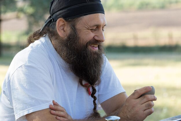 Um homem barbudo em uma bandana está bebendo chá no fundo da natureza.