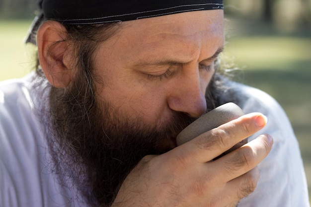 Um homem barbudo em uma bandana está bebendo chá no fundo da natureza.