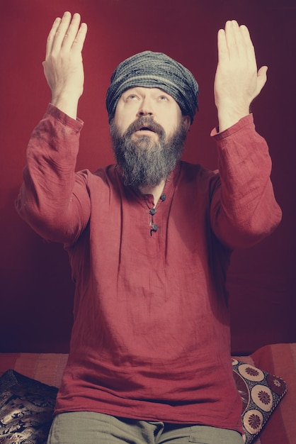 Foto um homem barbudo em uma camisa e um turbante em uma postura de oração levantou as mãos sobre um fundo vermelho toned