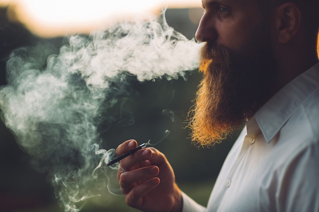 Um homem barbudo está fumando um cigarro contra o pôr do sol