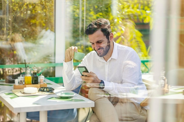 Um homem barbudo feliz em uma camisa com um sorriso no rosto está sentado no refeitório bebendo café da manhã fresco e rolando no telefone