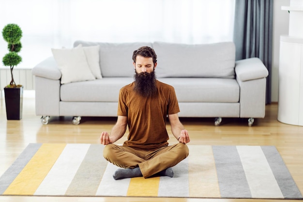 Um homem barbudo sentado no chão em sua casa aconchegante relaxando e meditando