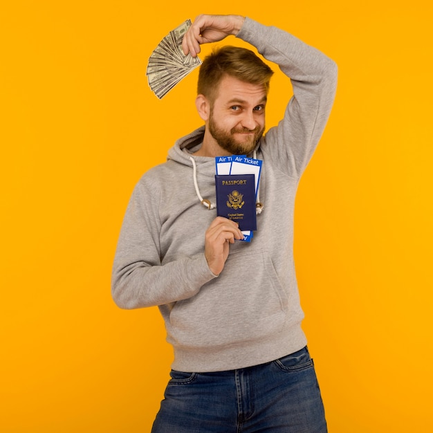 Um homem bonito com um moletom cinza se alegra em ganhar na loteria. ele está segurando um passaporte com passagens aéreas e dinheiro