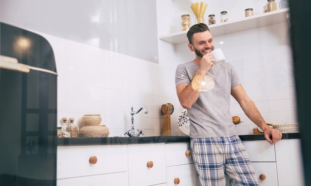 Um homem bonito de pijama está em pé com uma xícara em sua cozinha, bebendo alguma coisa e sorrindo.