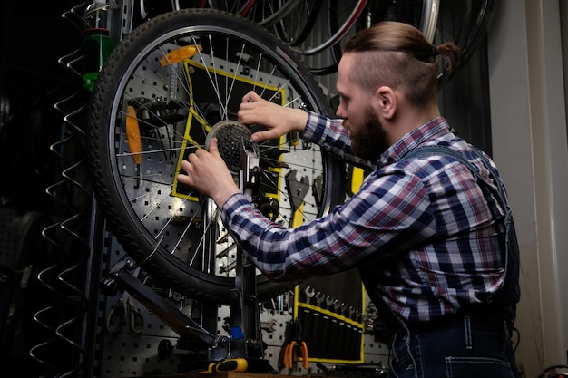 Um homem bonito e elegante vestindo uma camisa de flanela e macacão jeans, trabalhando com uma roda de bicicleta em uma oficina.