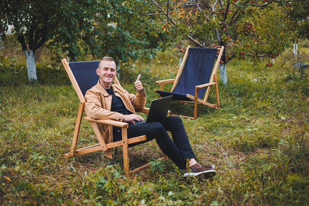 Um homem bonito em uma camisa marrom senta-se em uma cadeira dobrável de madeira no jardim e trabalha com um laptop Móveis ecológicos de madeira Trabalho remoto