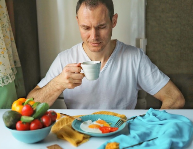 Um homem bonito em uma camiseta branca está sentado em uma mesa com um café da manhã saudável e bebendo café