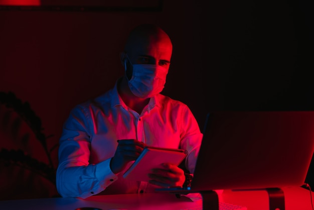 Um homem careca com uma máscara facial médica está trabalhando remotamente em um laptop em casa