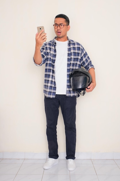 Foto um homem carregando um capacete de motocicleta mostrando uma expressão chocada ao olhar para seu telefone