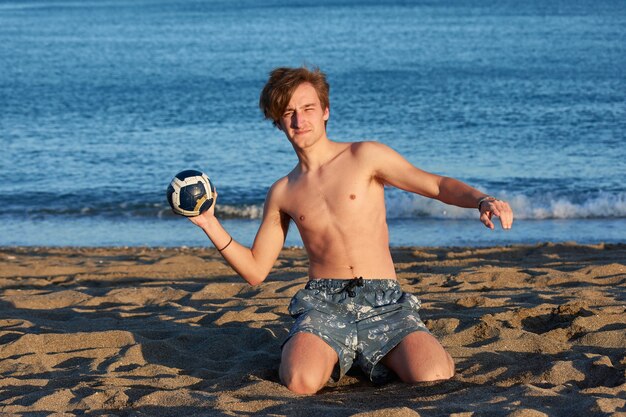 Um homem caucasiano brincando com uma bola em uma praia com o mar ao fundo