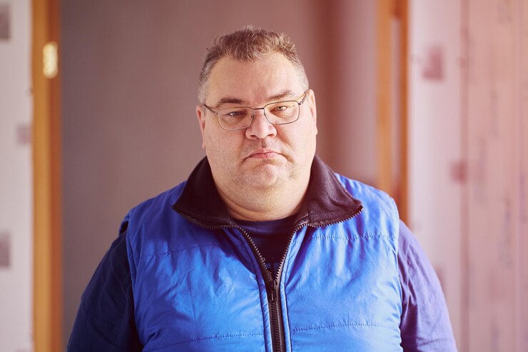 Foto um homem caucasiano com mais de 40 anos de idade, com óculos de prescrição no interior da casa, está no corredor.