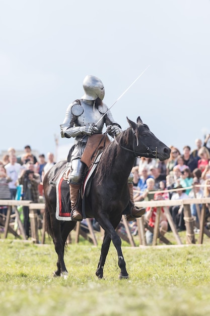 Um homem cavaleiro com uma espada andando a cavalo ao redor do campo de batalha pessoas assistindo atrás da cerca