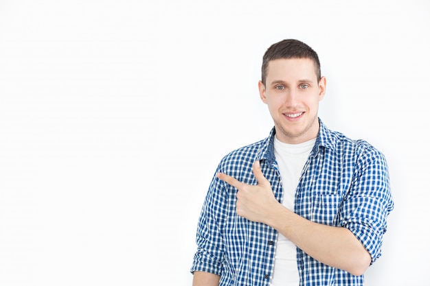 Um homem com a barba por fazer, estiloso, de camisa, aponta para uma cópia do espaço em uma parede branca, como mostra algo interessante, tem uma aparência sorridente, anuncia um produto. Olhe aqui! Pessoas, publicidade, conceito de emoção