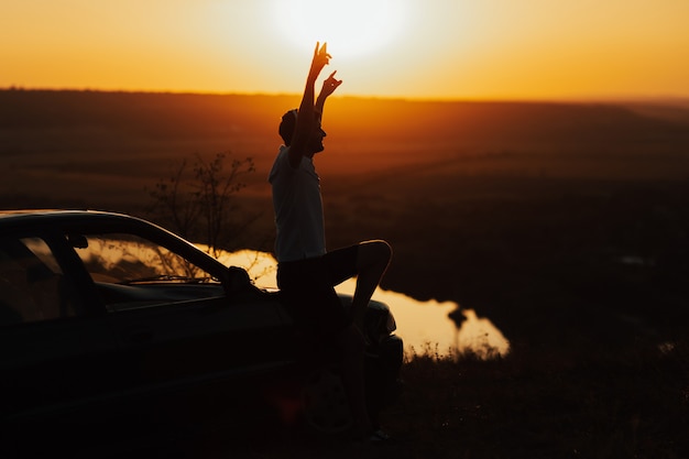 Um homem com as mãos ao alto admira uma vista costeira ao pôr do sol. homem sozinho no topo da montanha com o rio na superfície, sentado no capô do carro e aproveitando a viagem.