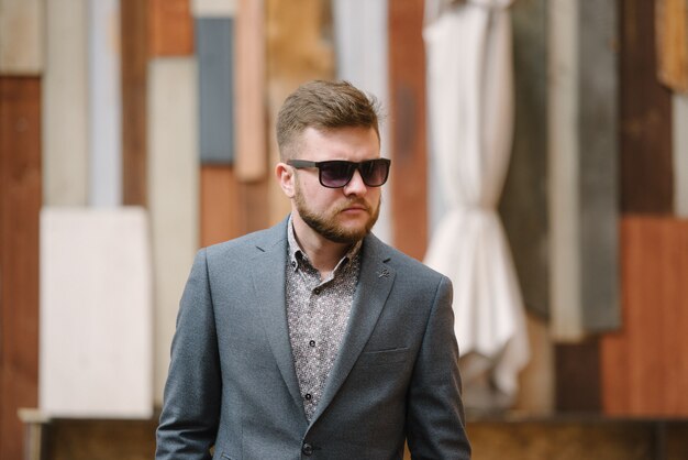 Um homem com barba e óculos escuros em um terno cinza posa na rua para anunciar roupas masculinas.