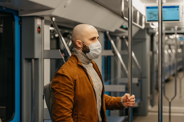 Um homem com barba e uma máscara médica para evitar a propagação do coronavírus está de pé e segurando o corrimão em um vagão do metrô. Um careca com máscara cirúrgica mantém distância social em um trem.