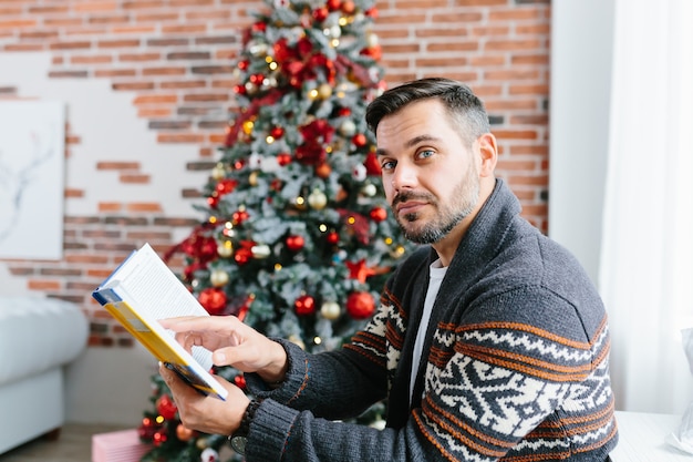 Um homem com barba em casa relaxando e lendo um livro, um empresário nos feriados de natal e ano novo em casa perto da árvore de natal