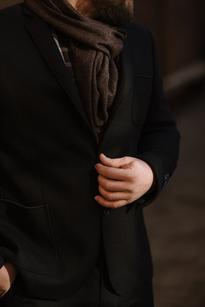 Um homem com barba em um casaco quente preto com um lenço posa para anunciar roupas masculinas no inverno. Anuncie roupas masculinas