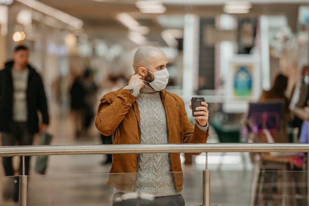 Um homem com barba está colocando uma máscara facial enquanto segura uma xícara de café marrom no shopping. Um careca mantém distância social.