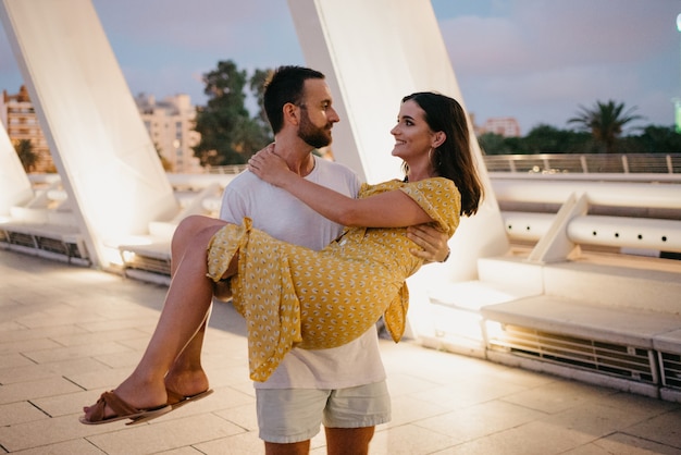 Um homem com barba está segurando sua namorada com um vestido amarelo nos braços em uma ponte em Valência. Um casal de turistas em um encontro na noite quente.