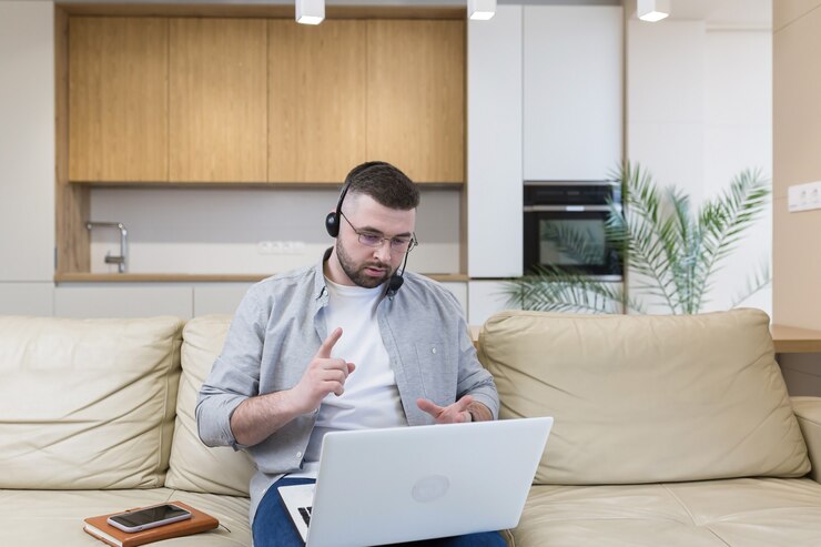 Foto um homem com barba que trabalha em casa sentado no sofá usa um laptop e um fone de ouvido com microfone realiza uma consulta online