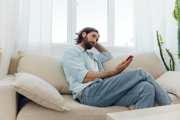 Um homem com barba senta-se no sofá durante o dia e sorri em casa e olha para o telefone relaxando em seu dia de folga online em seu telefone enviando mensagens de texto para amigos