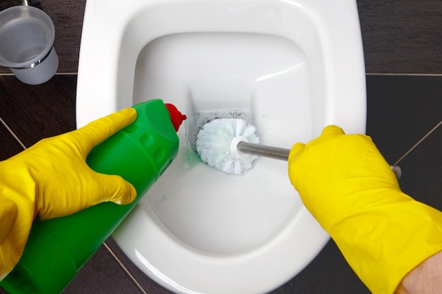 Um homem com luvas de borracha amarela limpa o banheiro com uma escova e um desinfetante