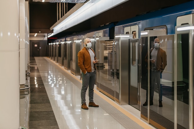 Um homem com máscara médica para evitar a disseminação do coronavírus segura um celular enquanto espera o trem do metrô. Um careca com máscara cirúrgica mantém distância social.