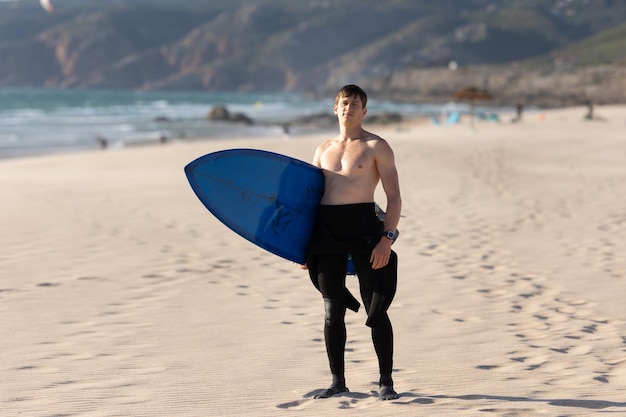 Um homem com torso nu em pé na praia segurando uma prancha de surf