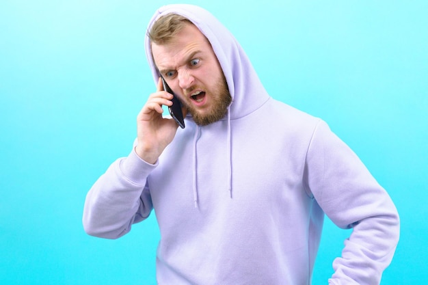 Um homem com um capuz roxo está louco gritando para seu telefone com a boca aberta em um fundo azul Pessoa Homem Surpresa Telefone Masculino Jovem Expressão Animado Cara Móvel Smartphone Retrato