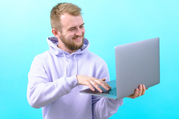 Um homem com um capuz roxo sorrindo e olhando para um laptop enquanto digita algum texto em um fundo azul Aprovação de sinal alegre Confiante Bom corporativo Bonito Alegria Positivo Bem-sucedido