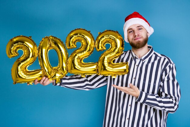 Foto um homem com um chapéu de natal segura os números 2022 em balões infláveis