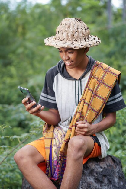 Foto um homem com um chapéu de palha está sentado em um tronco em um campo e olha para um telefone.